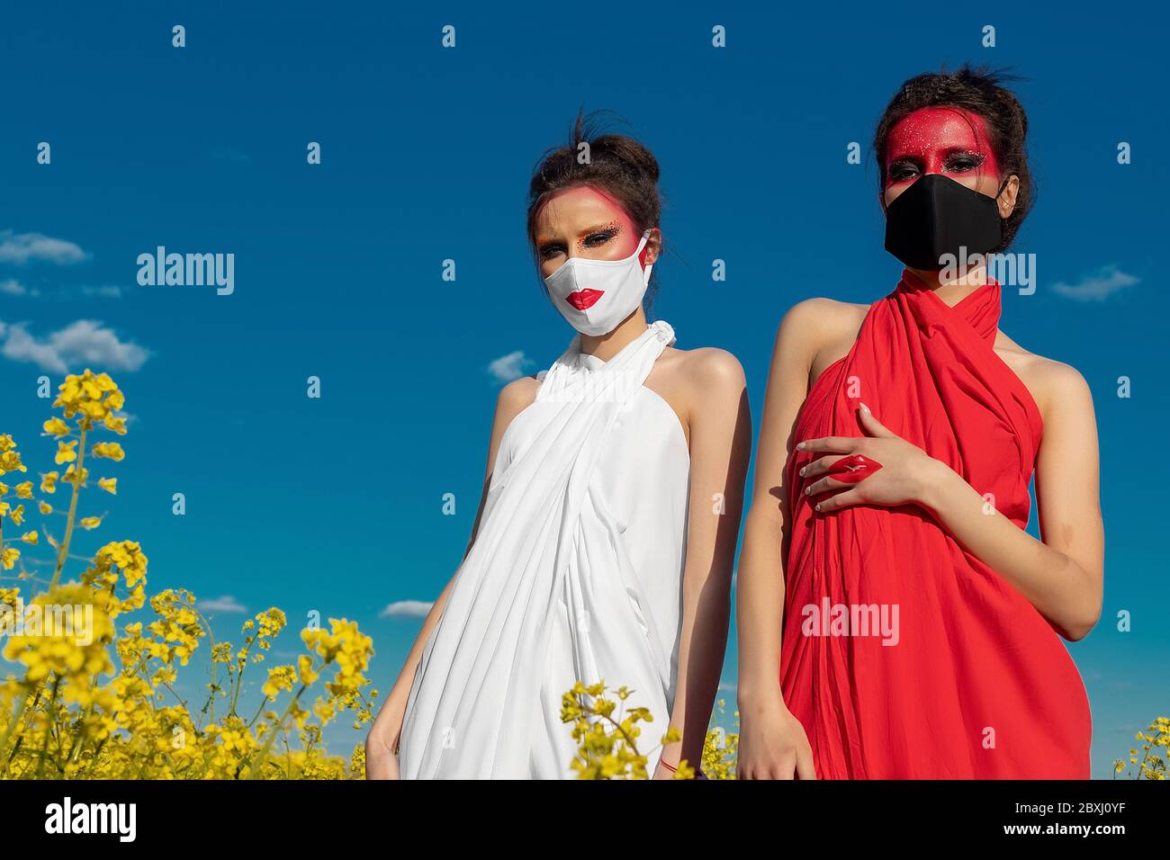 Two beautiful young brunette girls with creative bright makeup in tunics on a background of a field of yellow flowers and blue sky. One girl in a mask Stock Photo
