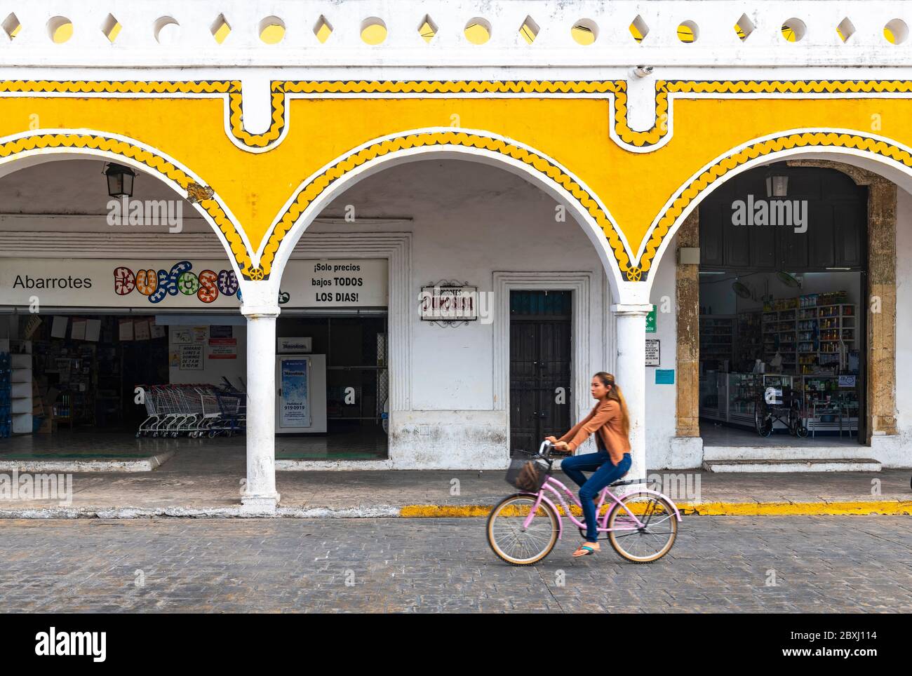 Young mexican woman on a bicycle with blur motion in the yellow city of Izamal with arch architecture by the main square, Yucatan Peninsula, Mexico. Stock Photo