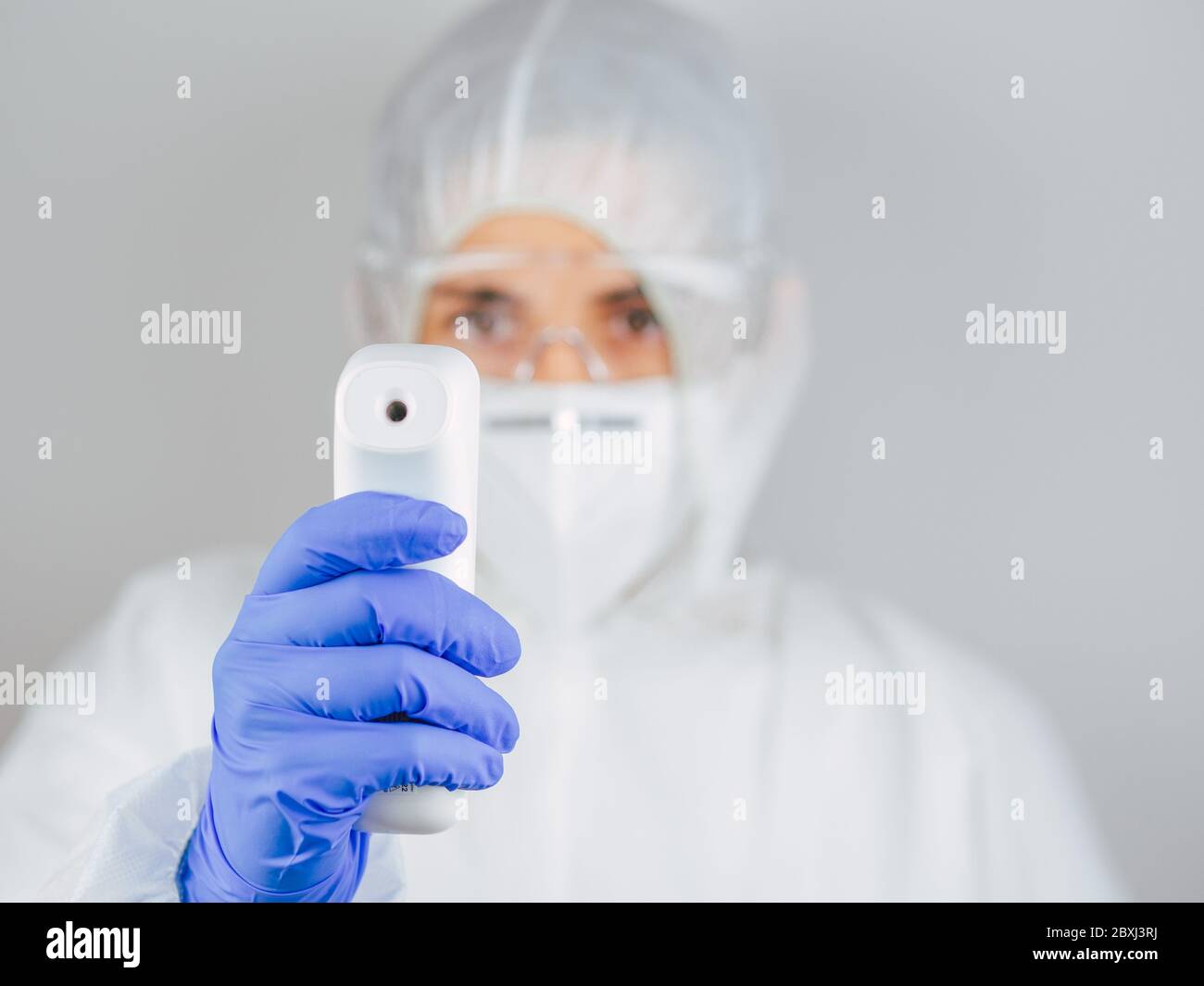 Close-up shot of female doctor wearing protective surgical mask, suit and glasses ready to use infrared forehead thermometer to check body temperature Stock Photo
