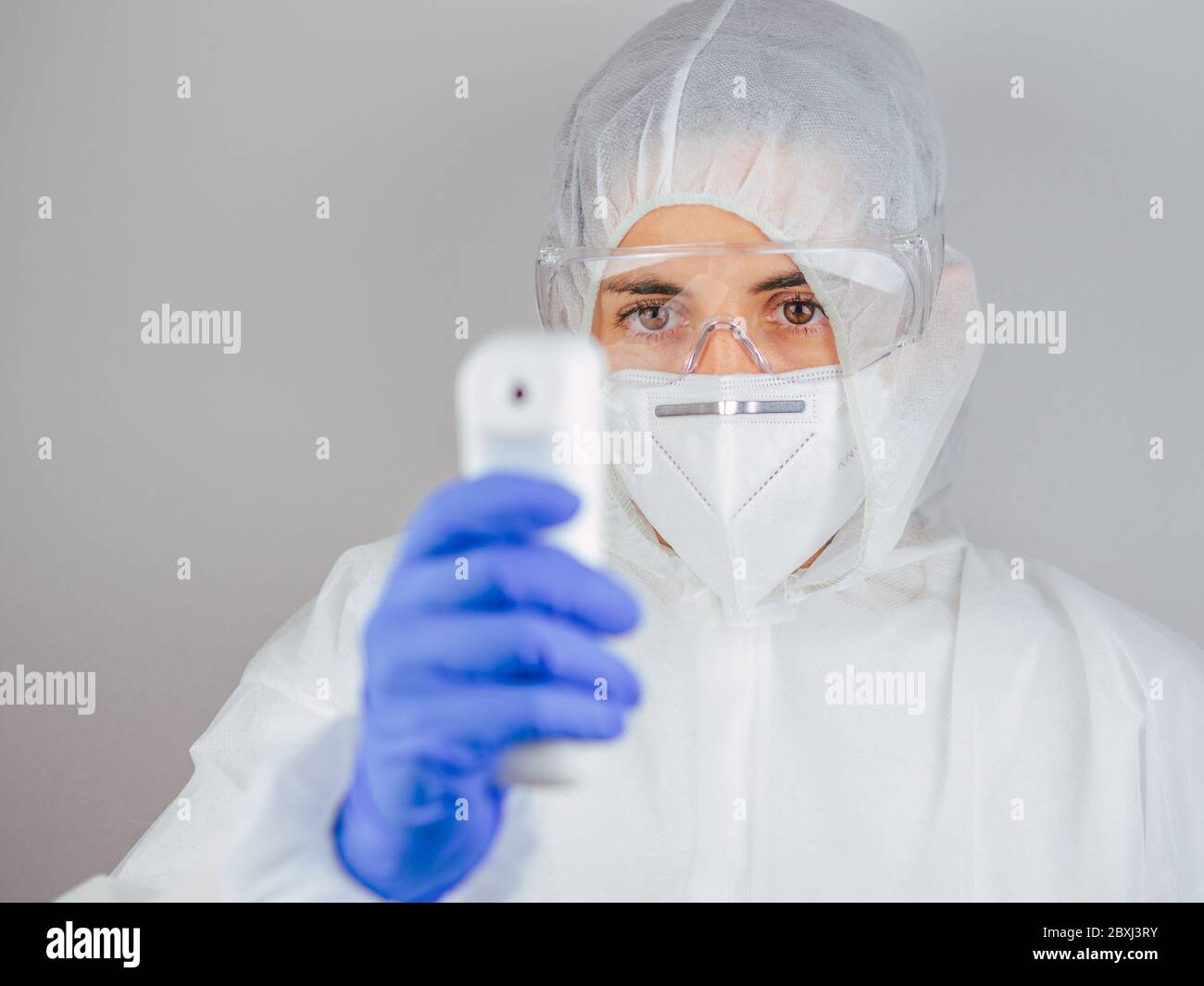 Close-up shot of female doctor wearing protective surgical mask, suit and glasses ready to use infrared forehead thermometer to check body temperature Stock Photo