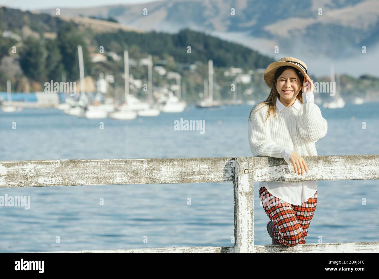 Beautyful asian woman resting is looks at the sea bay with houses and boats on a sunny day at Childrens bay, Akaroa, Canterbury, New Zealand. Relaxing Stock Photo