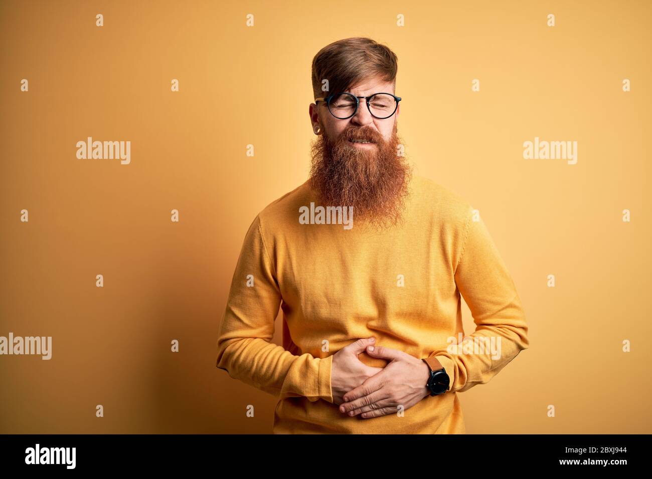 Handsome Irish redhead man with beard wearing glasses over yellow isolated background with hand on stomach because indigestion, painful illness feelin Stock Photo