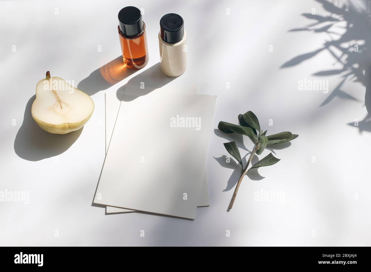 Summer stationery still life scene. Cut pear fruit, cosmetic oil, cream bottles and olive tree branch. White table. Blank paper cards, invitations moc Stock Photo