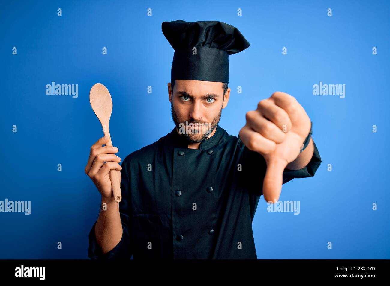 Young cooker man with beard wearing uniform holding wooden spoon over ...