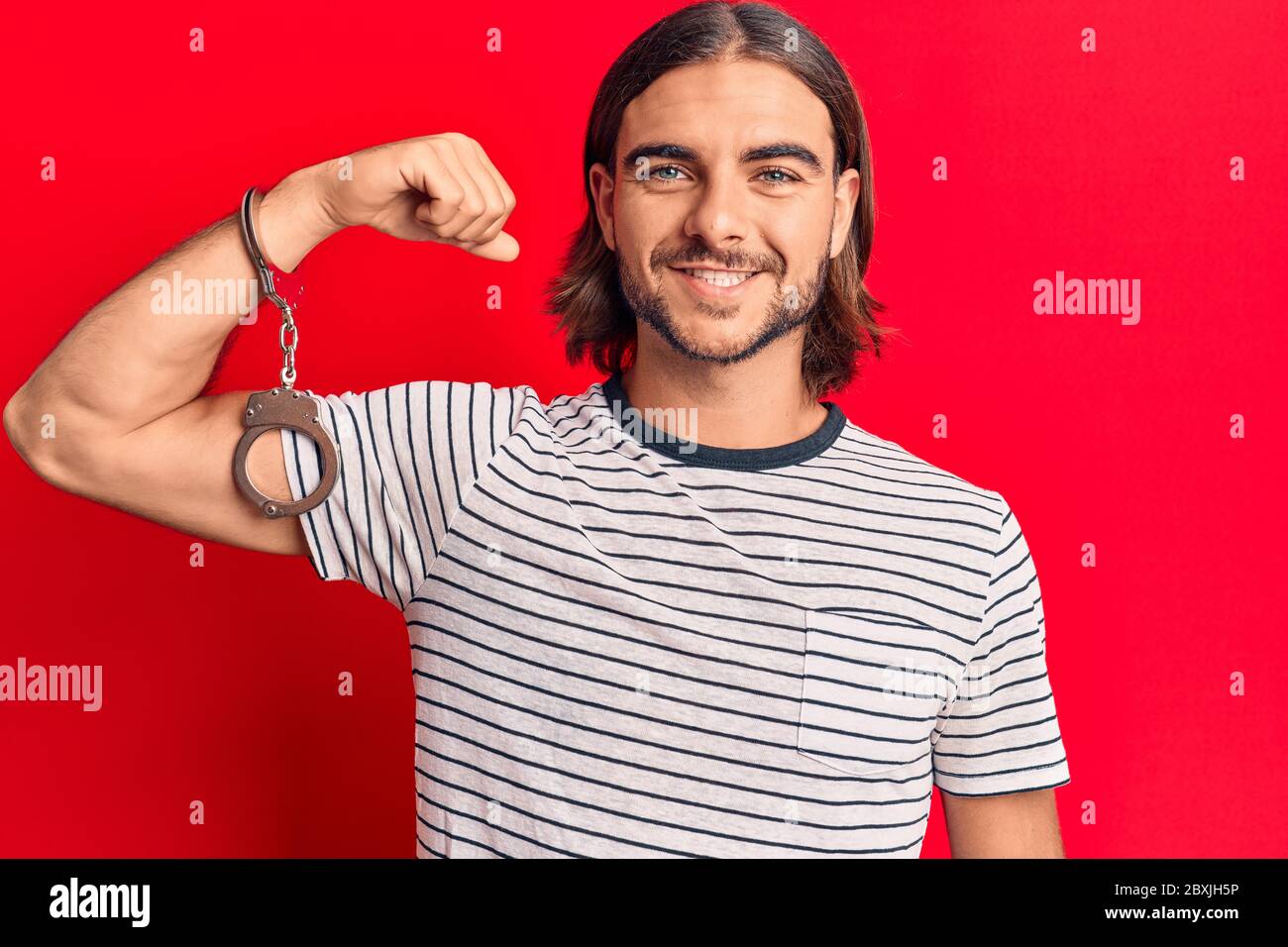 Young handsome man wearing prisoner handcuffs looking positive and happy standing and smiling with a confident smile showing teeth Stock Photo