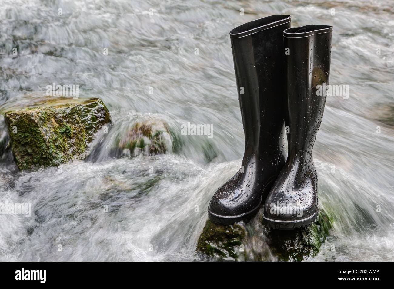 Black rubber boots stand on a stone in the rushing water of a creek.In wet and damp terrain you always have dry feet with rubber boots. Stock Photo