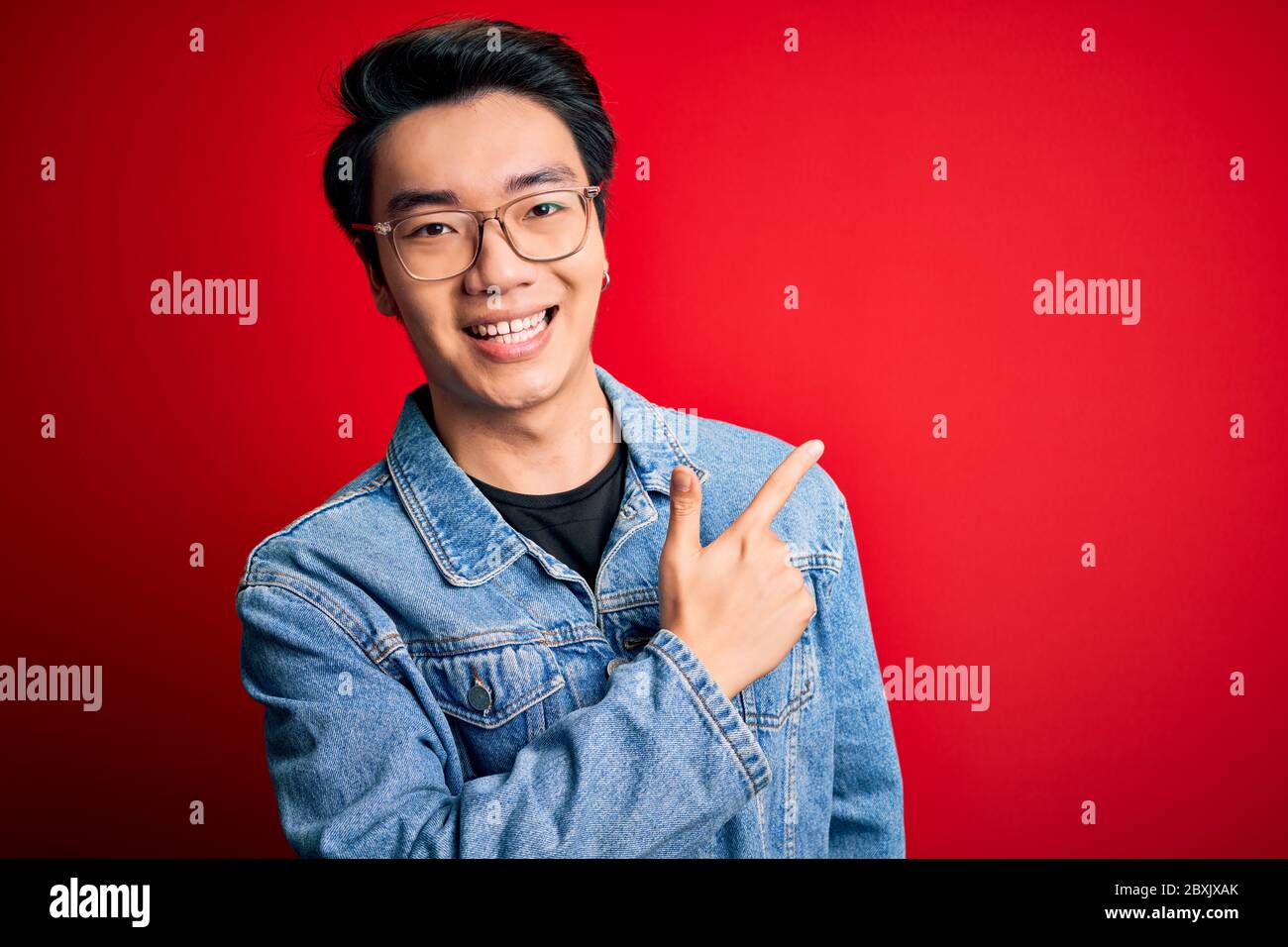 Young handsome chinese man wearing denim jacket and glasses over red