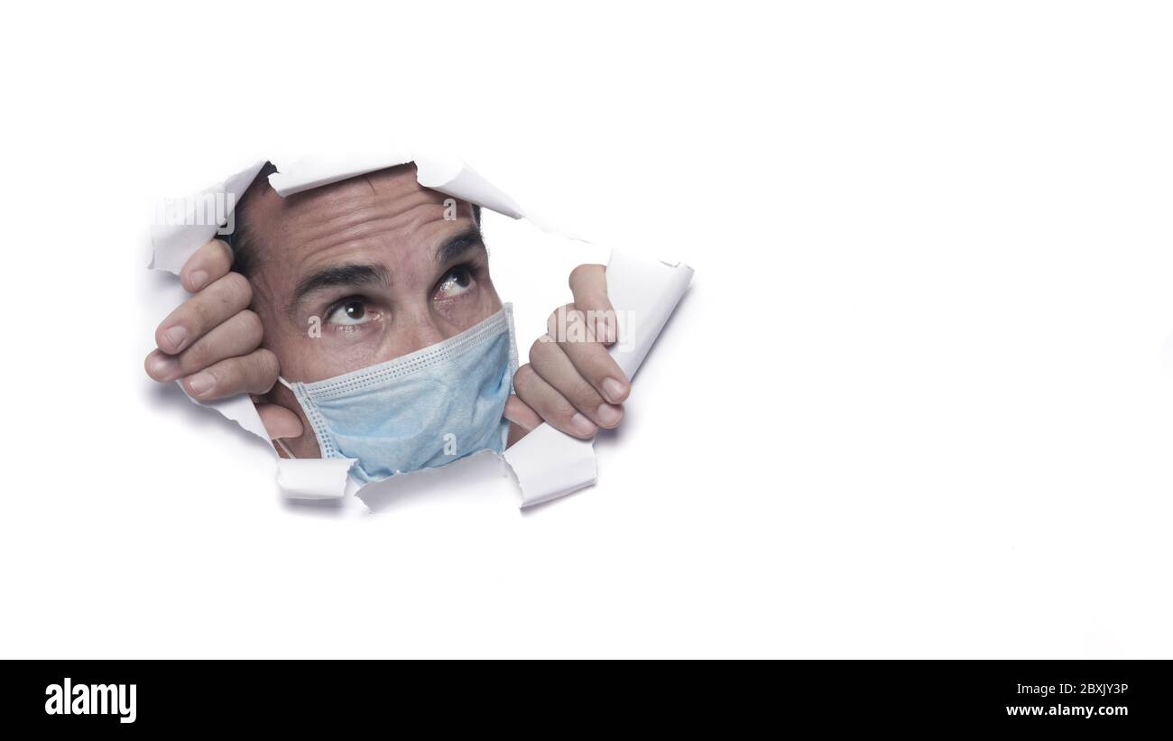 mid age caucasian man wearing protective mask or surgeon breaking through a white paper sheet or wall watching outside in confinement for coronavirus Stock Photo