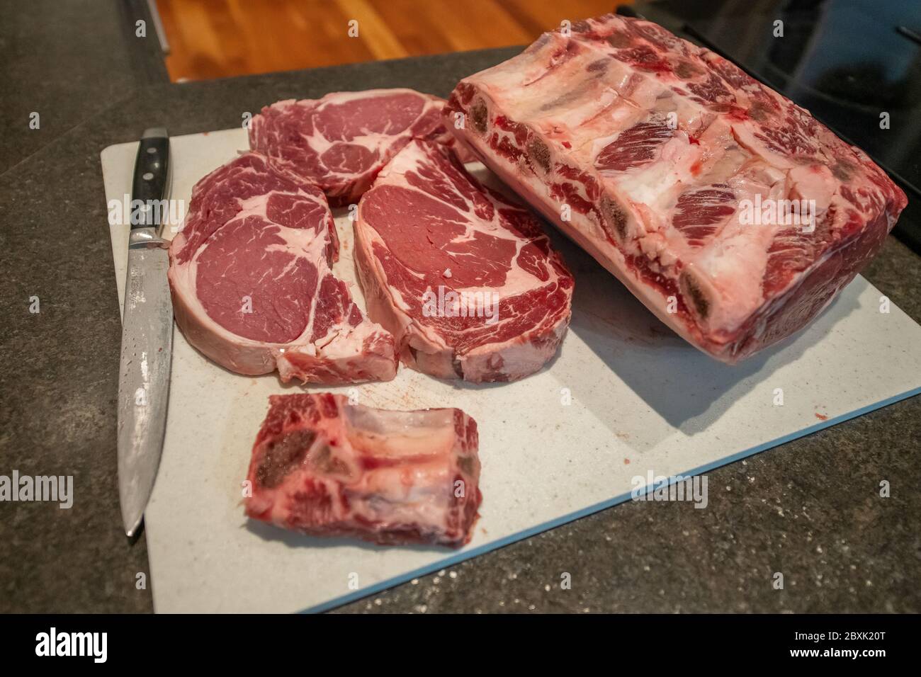 Raw beef prime rib roasts and steaks with thick pieces of meat, the marbling of fat and rib bones. The meat is sitting on a white cutting board. Stock Photo
