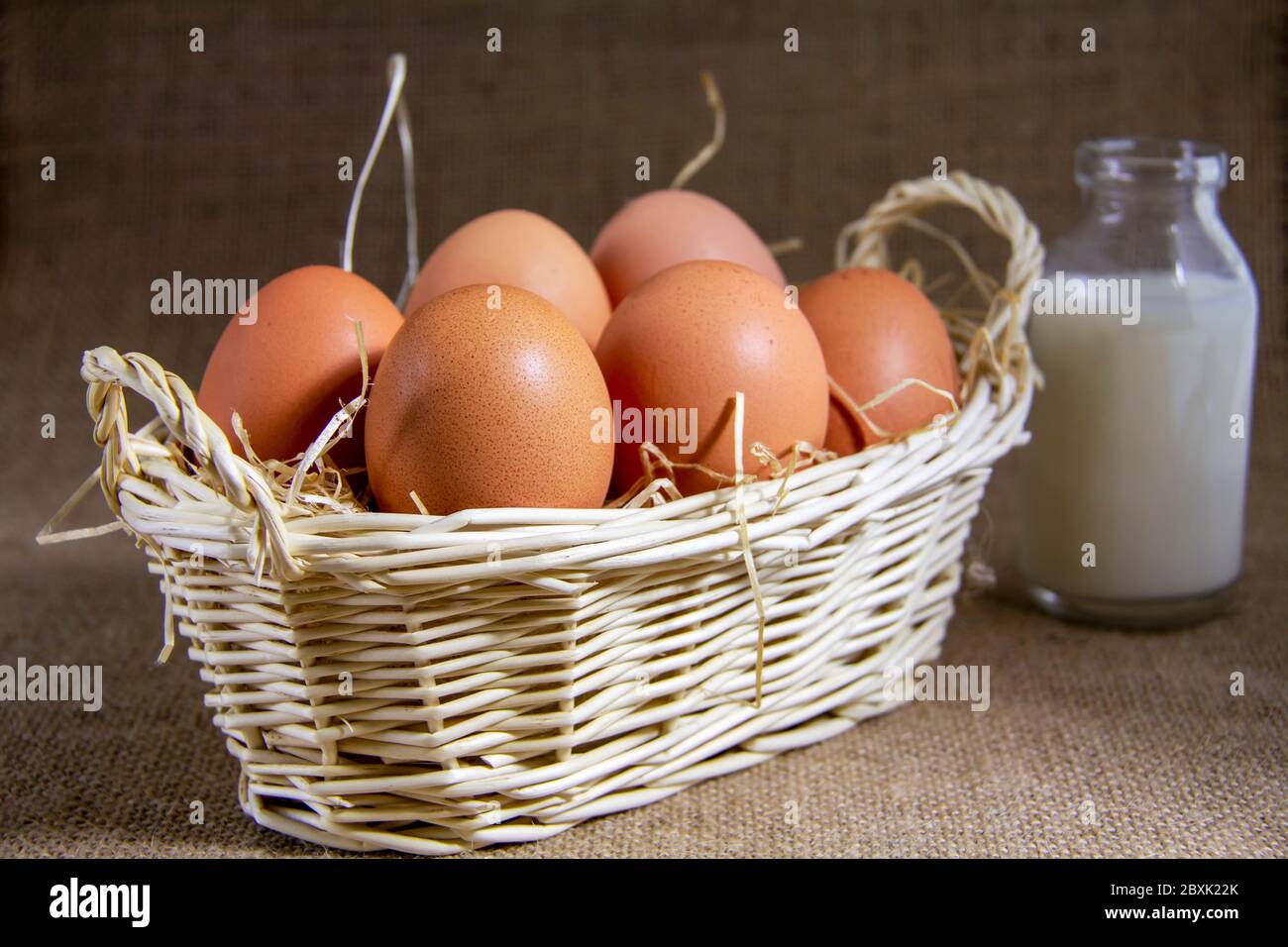 Six Eggs on some straw in a Wicker Basket, with a bottle of milk behind Stock Photo