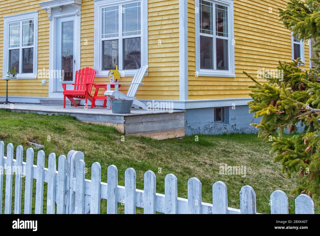 Corner view of a vintage yellow wooden clapboard house with red and white adirondack chairs on the patio. The building has multiple windows. Stock Photo