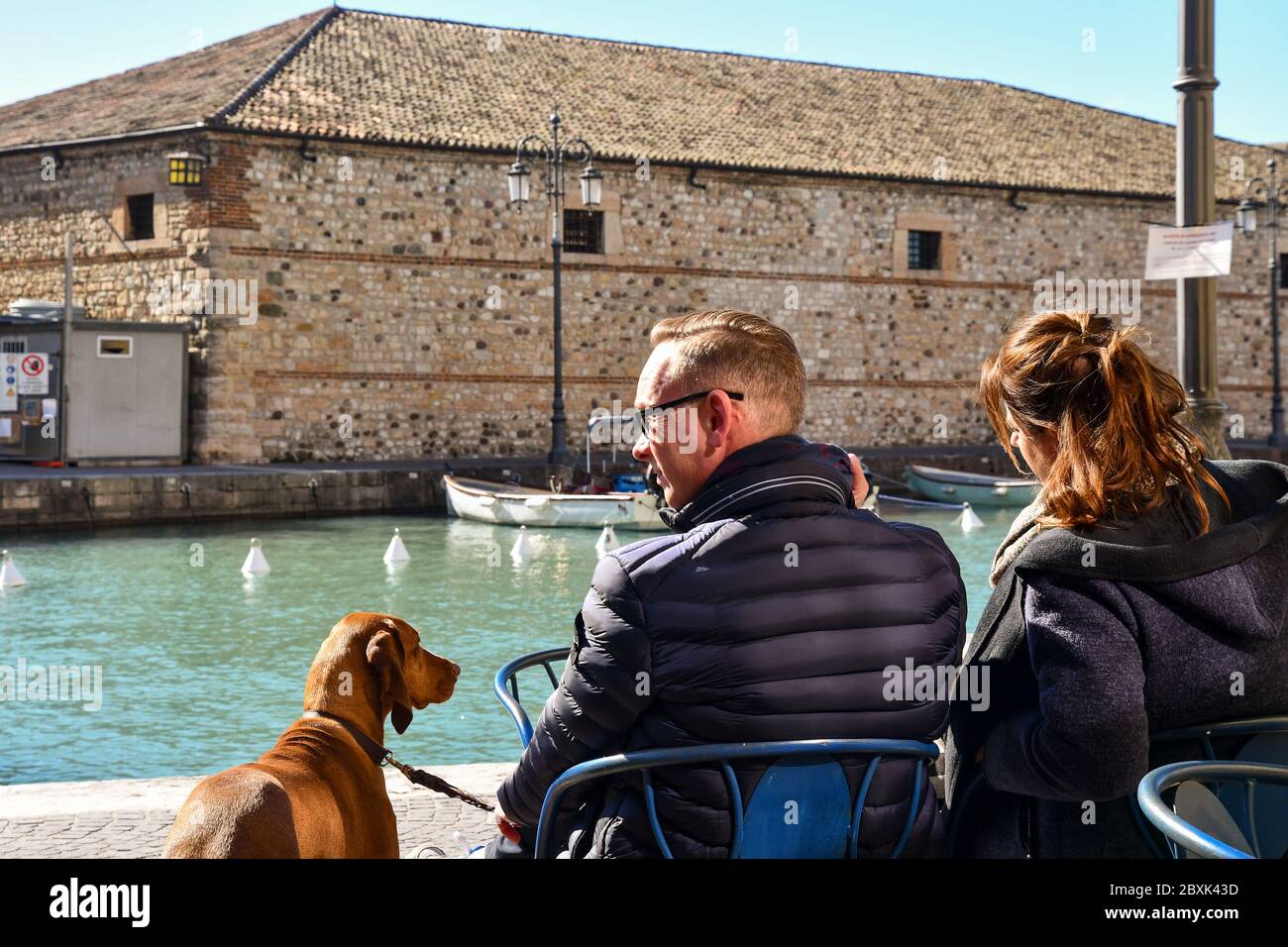 A couple with their dog in an outdoor cafe overlooking the canal, with the Venetian Customhouse in the background, Lazise, Verona, Veneto, Italy Stock Photo