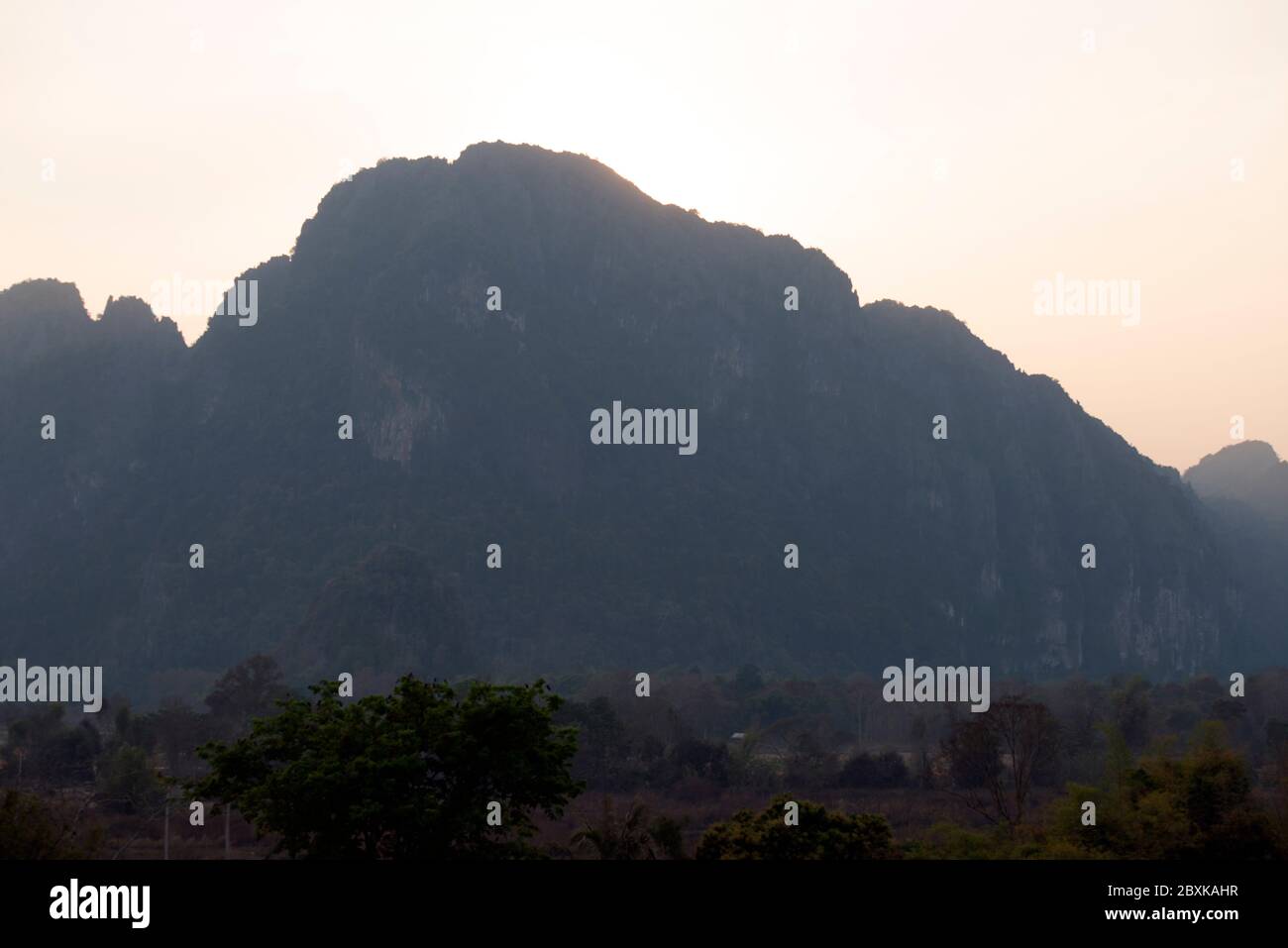 The sun sets behind a limestone karst in A hot air balloon hovers in Vang Vieng, Laos, Southeast Asia. Stock Photo