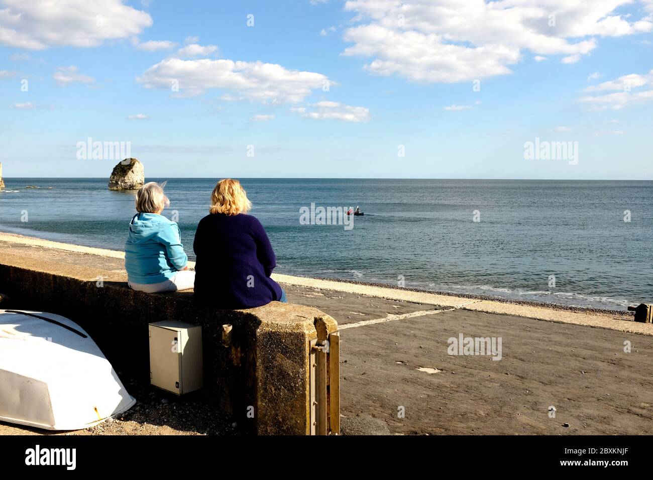 Two women sitting chatting socialising on the promenade wall of Freshwater bay on the Isle of Wight Stock Photo
