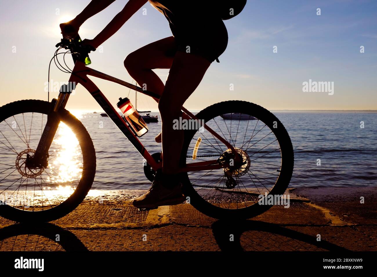 Cycling along seafront sea wall promenade revetment as sun setting over horizon in background shining through wheel rider in silhouette Stock Photo