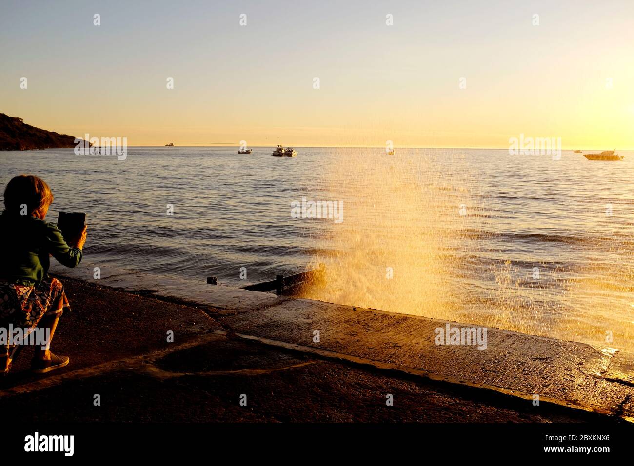 Woman taking a photo using her mobile phone of a wave crashing onto the sea wall revetment and sunset sunlight shining through the spray orange glow Stock Photo