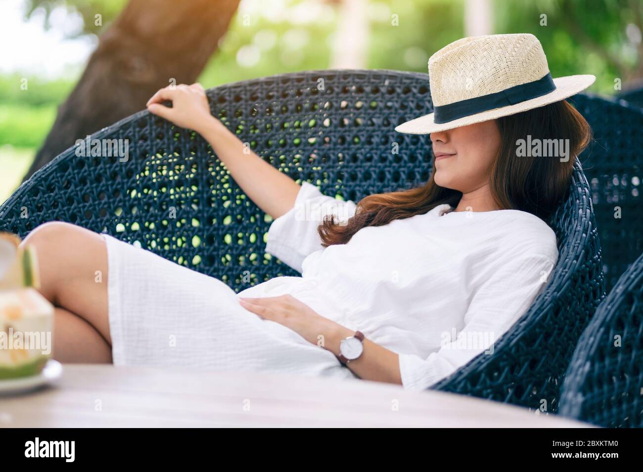Portrait image of a beautiful asian woman with hat lying down on a bench in the garden with coconut on the table Stock Photo