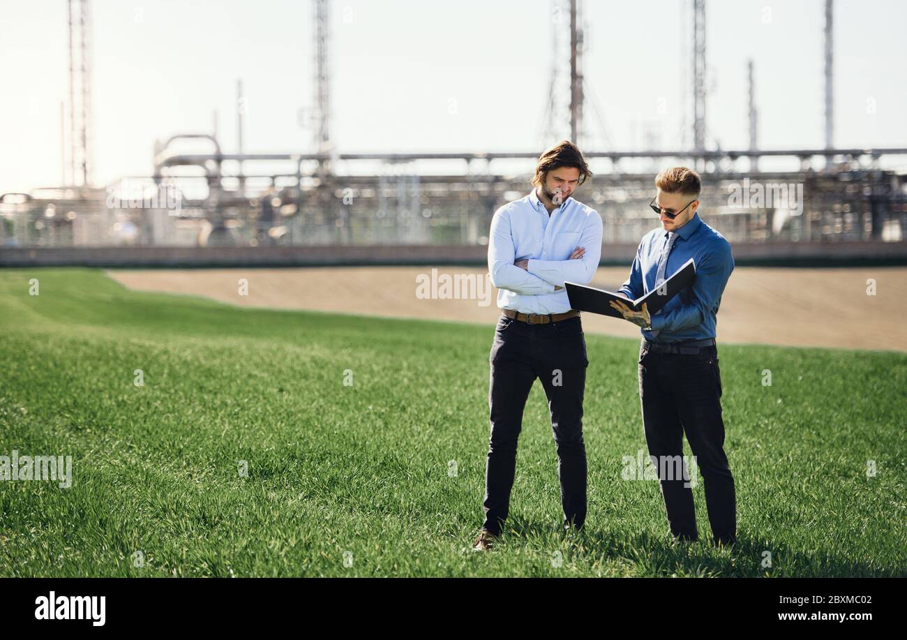 Two young engineers standing outdoors by oil refinery, discussing issues. Stock Photo