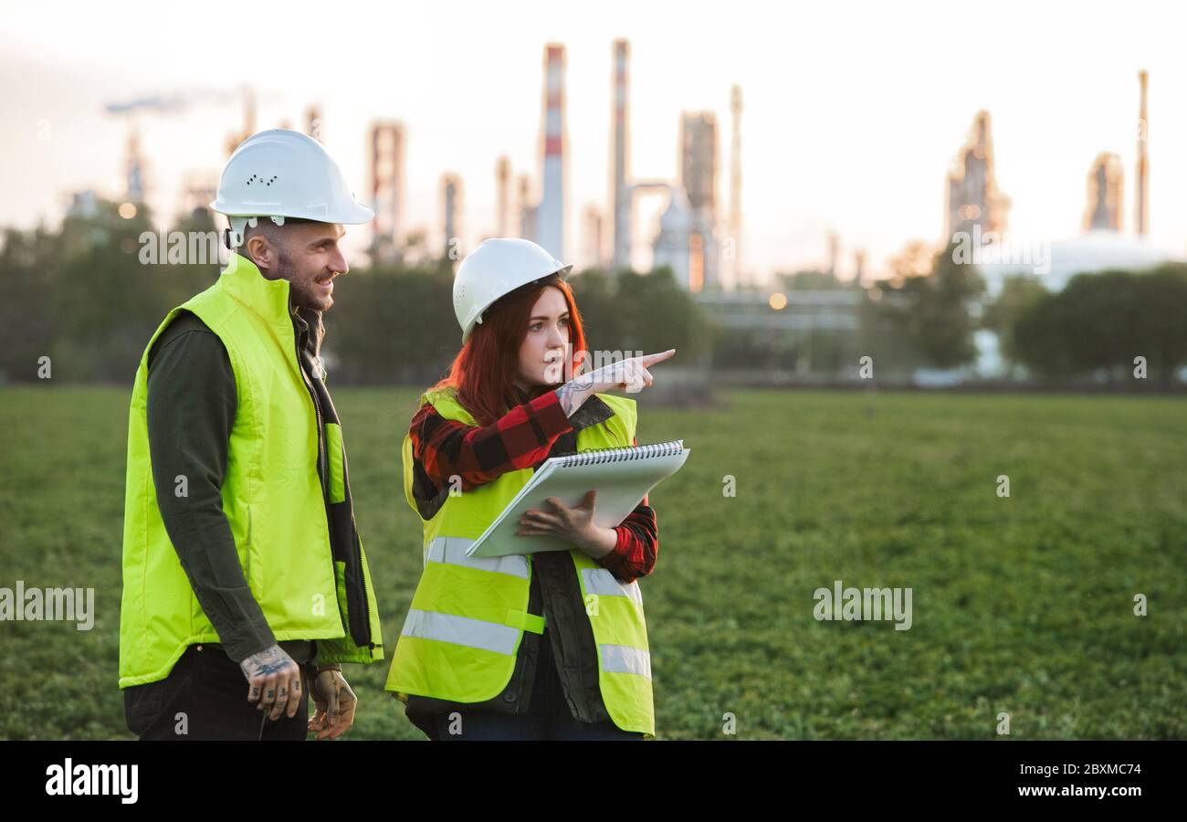 Two young engineers standing outdoors by oil refinery, discussing issues. Stock Photo