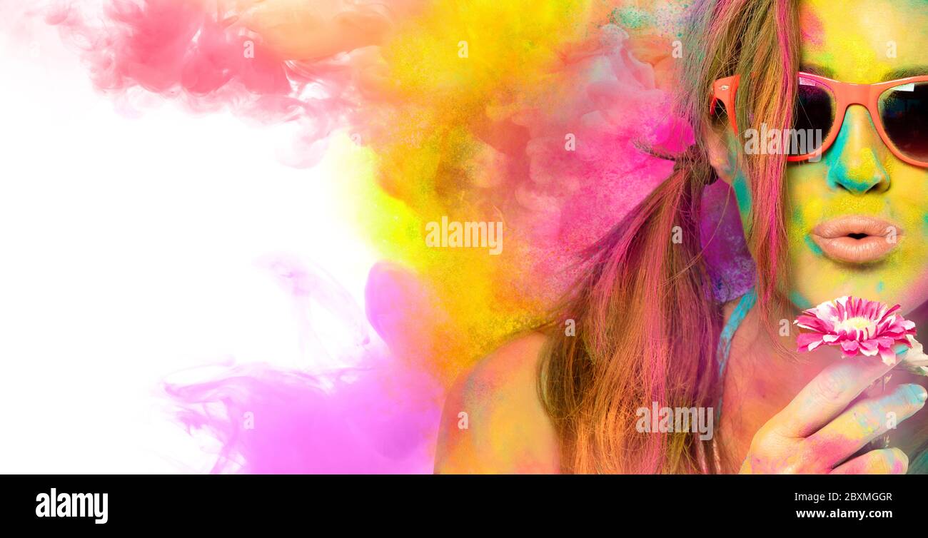 Beautiful young woman wearing stylish sunglasses blowing on a flower celebrating the Holi festival covered in brightly colored powder with clouds of c Stock Photo