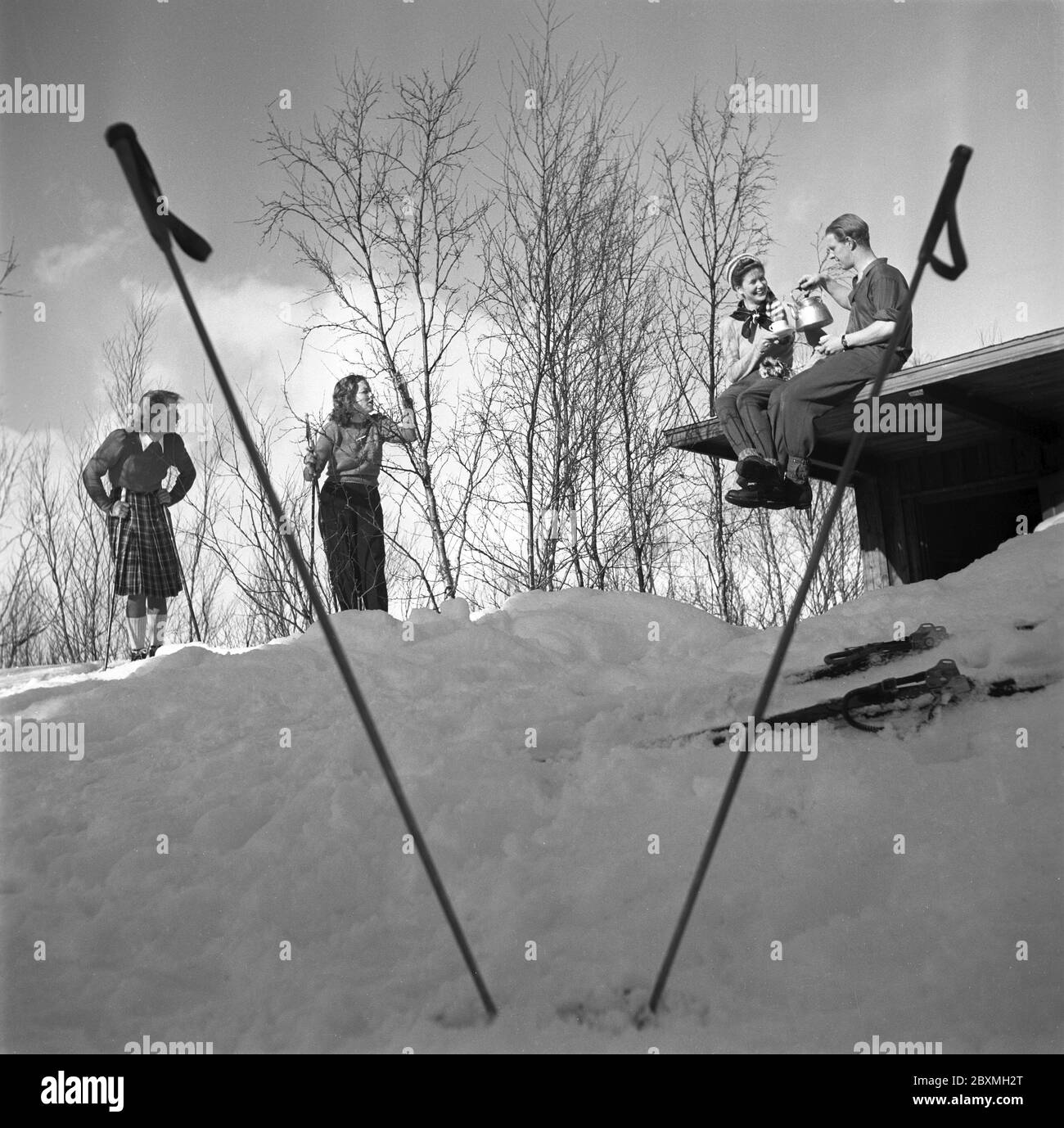 Winter in the 1940s. A young couple is having a cup of coffee while sitting on the roof of their cottage. Their two friends are standing on skis and chatting. The picture is taken in Swedish Lapland near Björkliden. It's Mr and Mrs Iris and Bertil Thorelli having a nice time in the Swedish wilderness.   Sweden 1943 Photo Kristoffersson Ref D115-1 Stock Photo