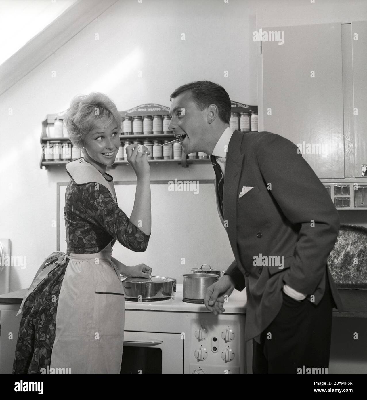 In the kitchen 1950s. A young couple in their kitchen. She is making him taste the food and puts a spoon in his mouth. Sweden 1959 Kristoffersson ref CG35 Stock Photo