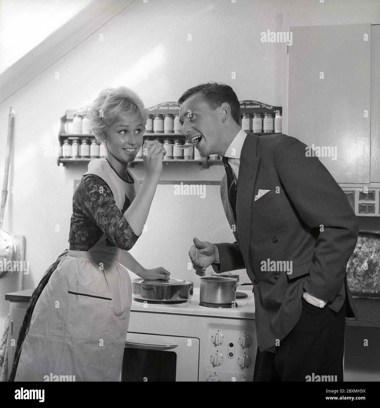 In the kitchen 1950s. A young couple in their kitchen. She is making him taste the food and puts a spoon in his mouth. Sweden 1959 Kristoffersson ref CG35 Stock Photo