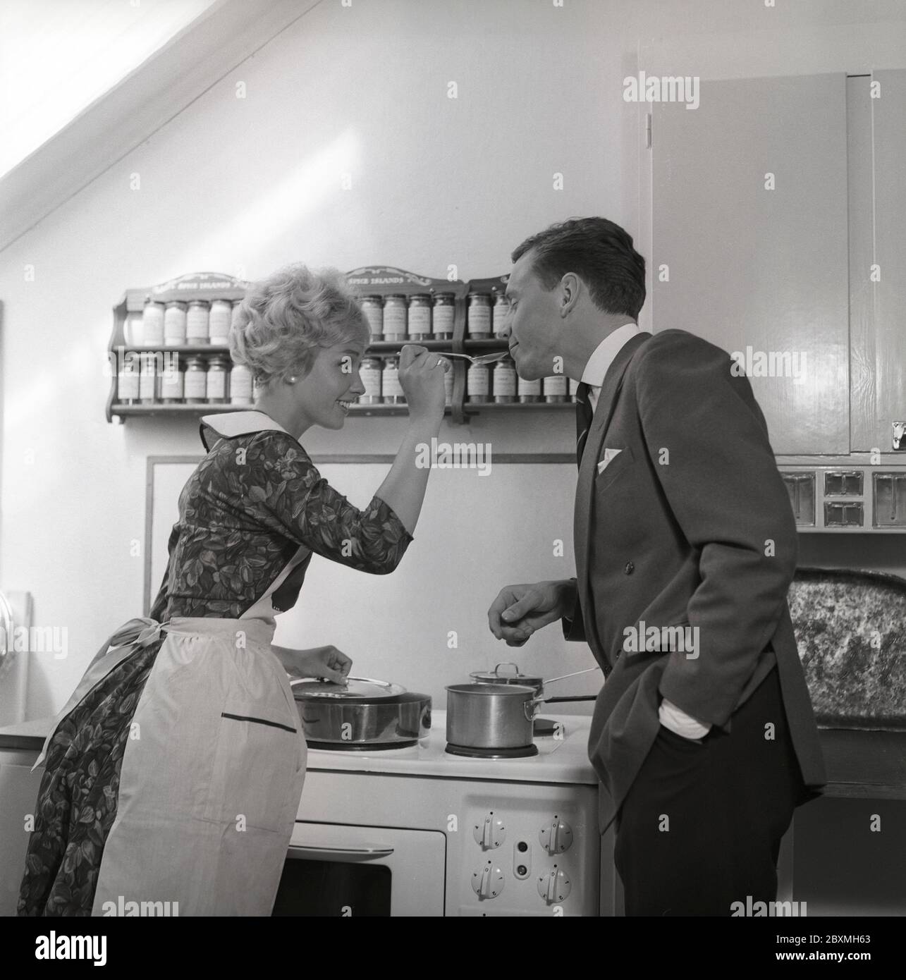 In the kitchen 1950s. A young couple in their kitchen. She is making him taste the food and puts a spoon in his mouth. Sweden 1959 Kristoffersson ref CG35 Stock Photo