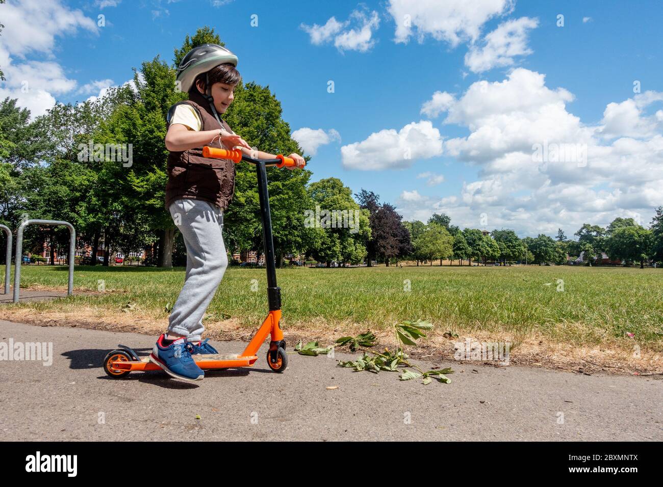 A young boy plays with and pushes his scooter on a sunny day in the park. Blue sky and green trees. Energetic and active child. Stock Photo