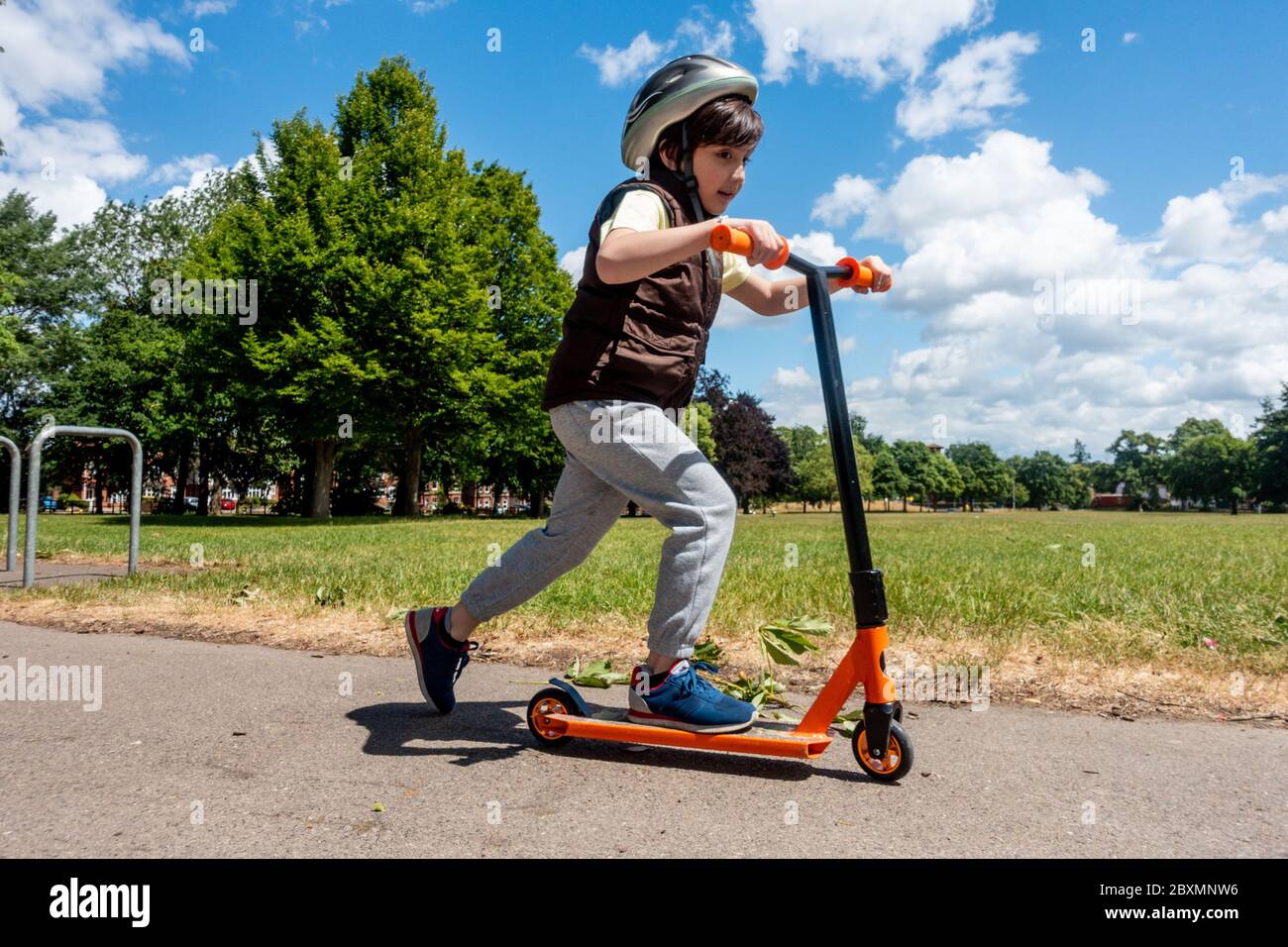 A young boy plays with and pushes his scooter on a sunny day in the park. Blue sky and green trees. Energetic and active child. Stock Photo