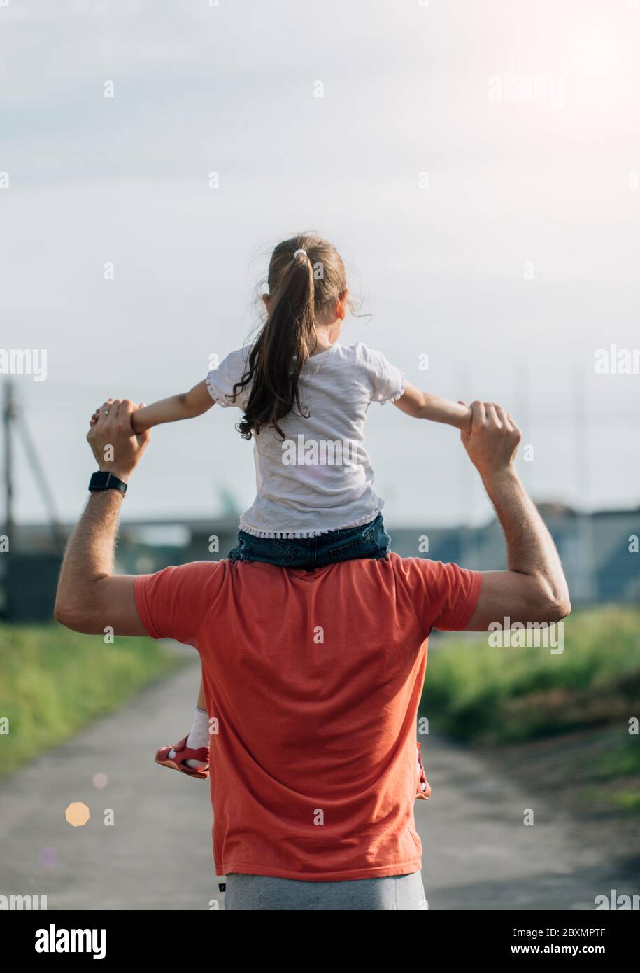Dad dancing on his shoulders with his daughter in sun. Father travels with baby on his shoulders in rays of sunset. Child with parents walks at sunset Stock Photo
