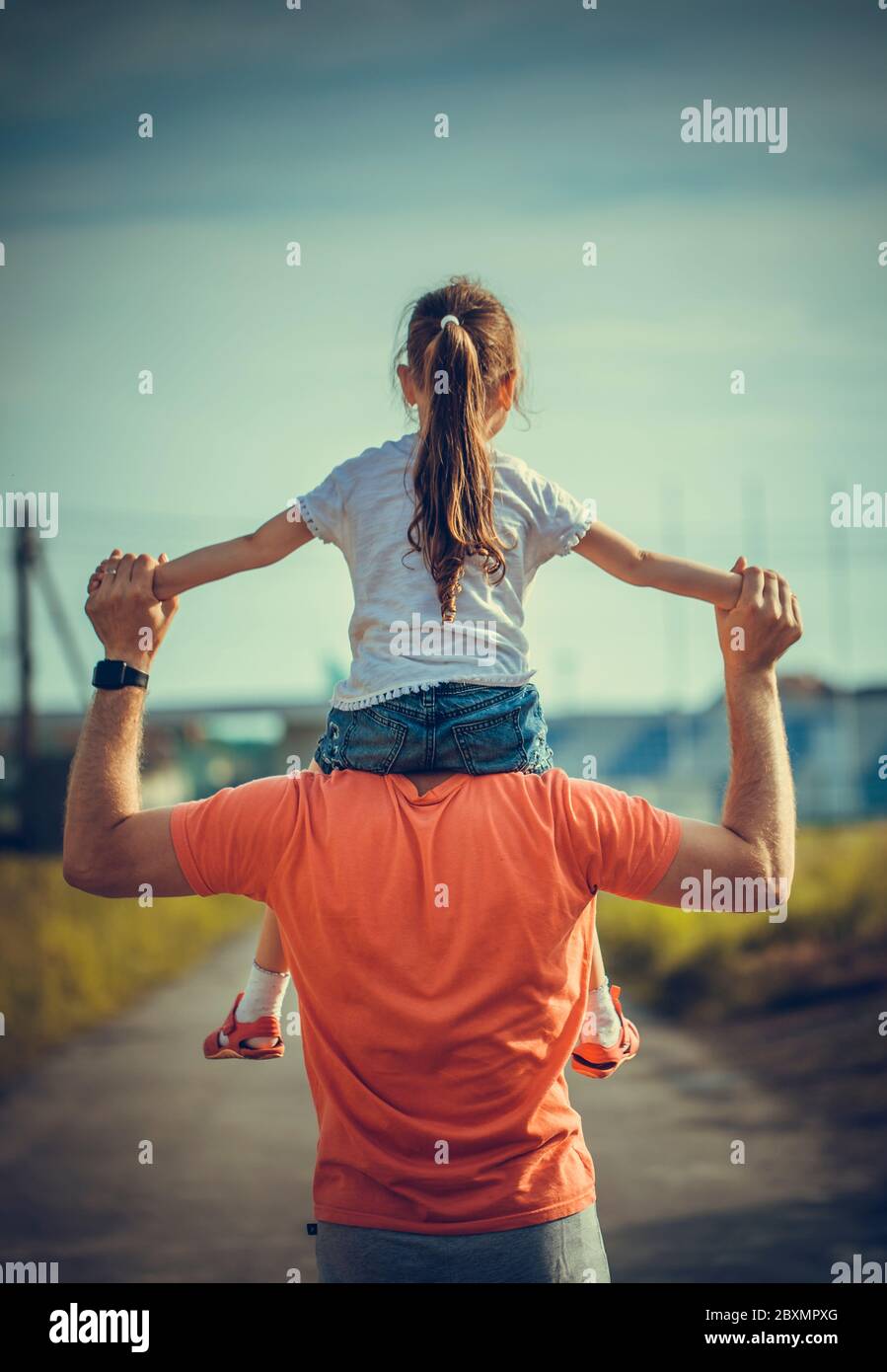 Dad dancing on his shoulders with his daughter in sun. Father travels with baby on his shoulders in rays of sunset. Child with parents walks at sunset Stock Photo