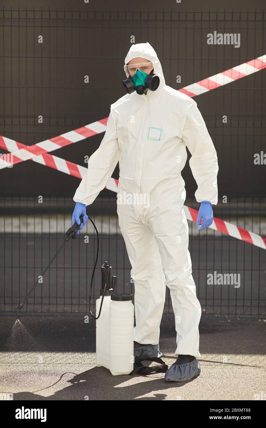 Vertical full length portrait of male worker wearing hazmat suit holding disinfection gear while standing outdoors with red caution tape in background Stock Photo