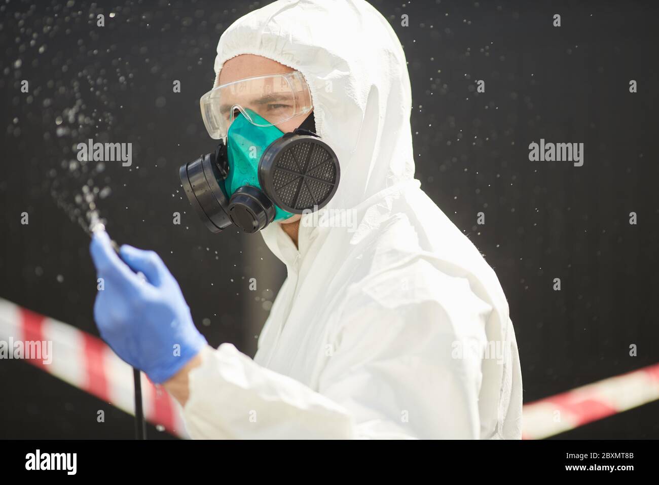 Chest up portrait of male worker wearing hazmat suit holding disinfection gear while standing outdoors with red caution tape in background Stock Photo