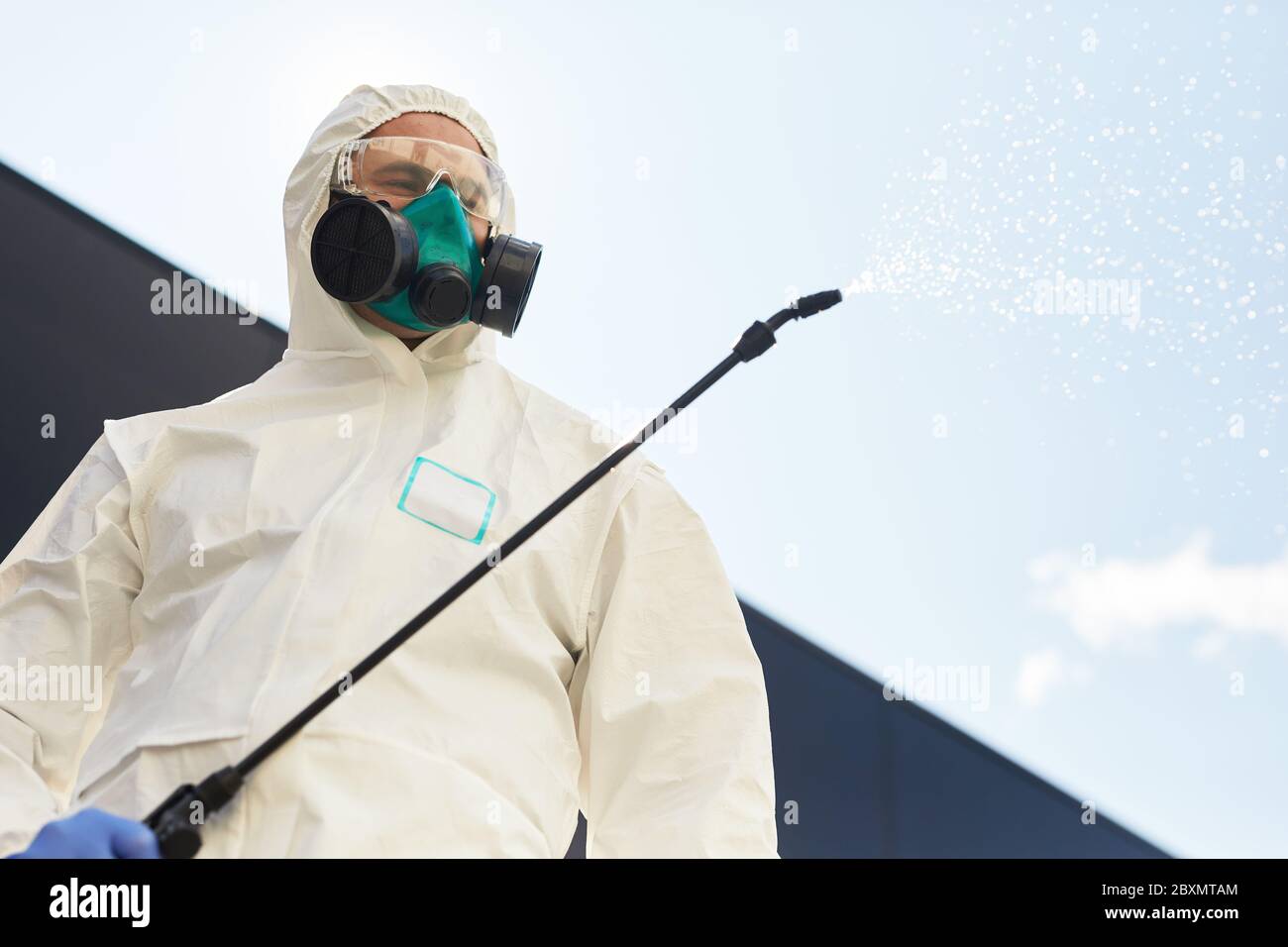 Low angle portrait of male worker wearing hazmat suit holding disinfection gear spraying chemicals outdoors while standing against sky background, cop Stock Photo