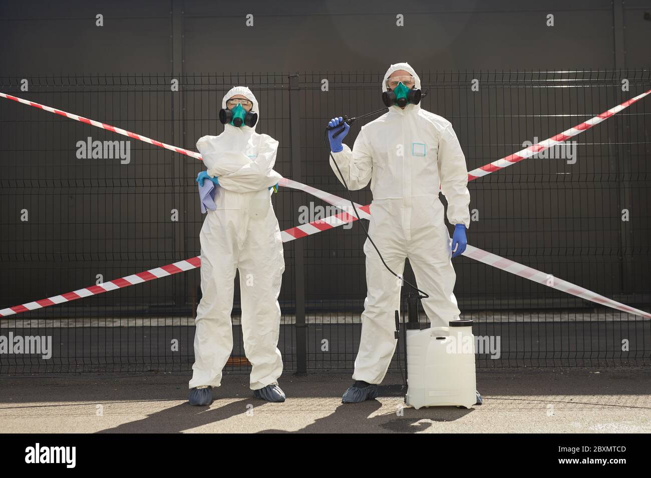 Full length portrait of two workers wearing hazmat suits holding disinfection gear while standing outdoors with red caution tape in background Stock Photo