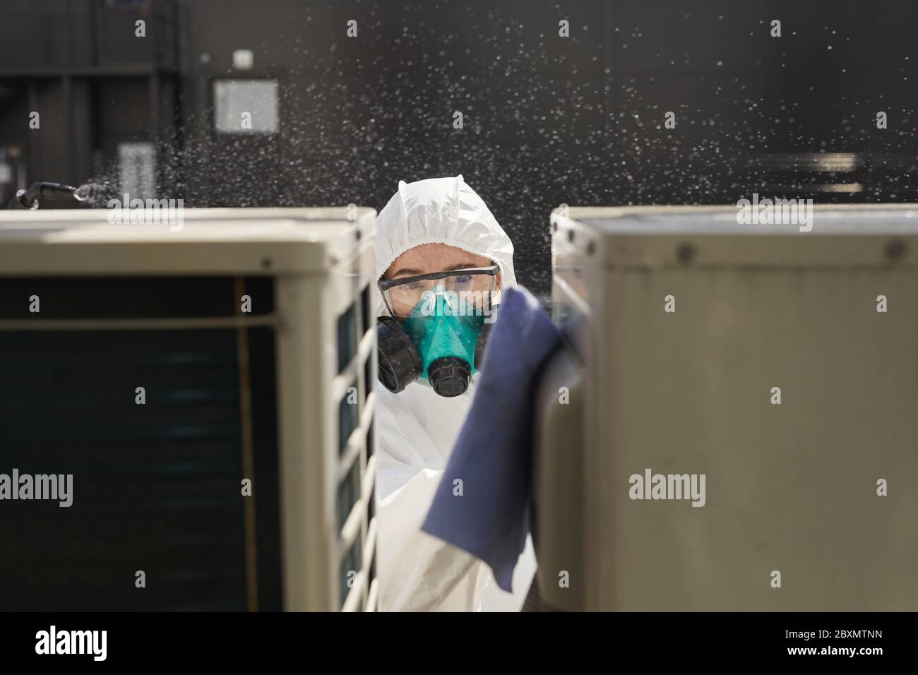 Portrait of female worker wearing protective suit and respirator disinfecting surfaces outdoors lit by sunlight, copy space Stock Photo