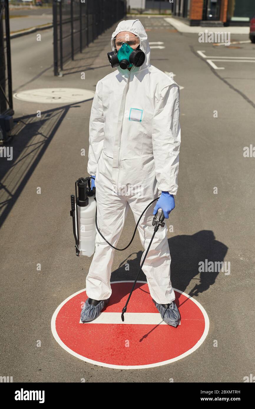 Vertical full length portrait of male worker wearing hazmat suit and holding disinfecting gear while standing on STOP sign outdoors lit by sunlight Stock Photo