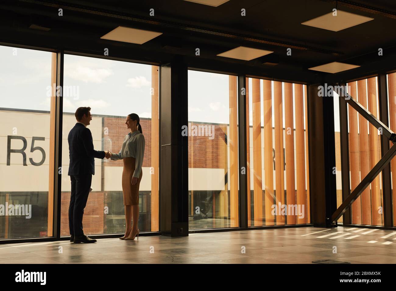 Wide angle view of real estate agent shaking hands with client while standing in empty office building interior lit by sunlight, copy space Stock Photo