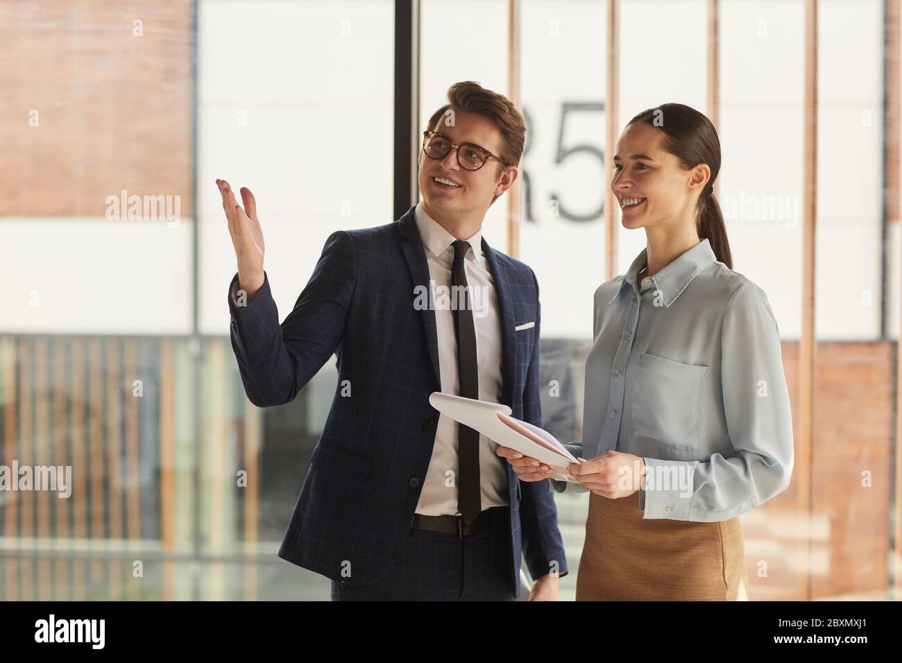 Waist up portrait of smiling real estate agent discussing property with female client and pointing up while standing in empty office building interior Stock Photo