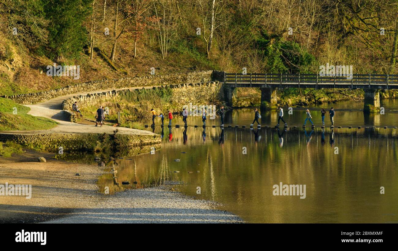 People walking on stepping-stones across water of River Wharfe (by footbridge) in sunshine - scenic Bolton Abbey estate, Yorkshire Dales, England, UK. Stock Photo