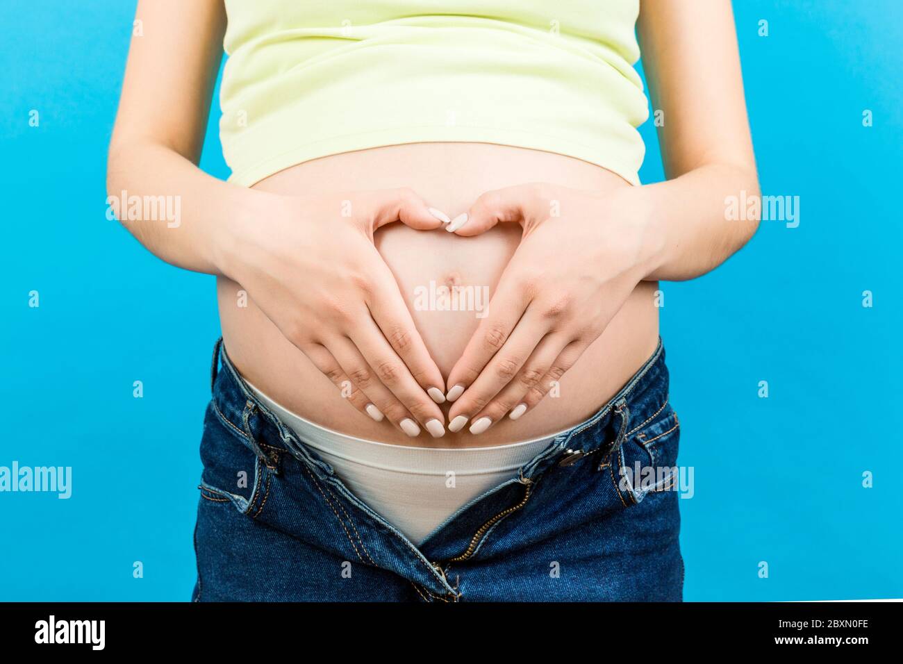 Close Up Of Pregnant Woman In Unzipped Jeans Holding Her Hands In A Heart Shape On Her Belly At 