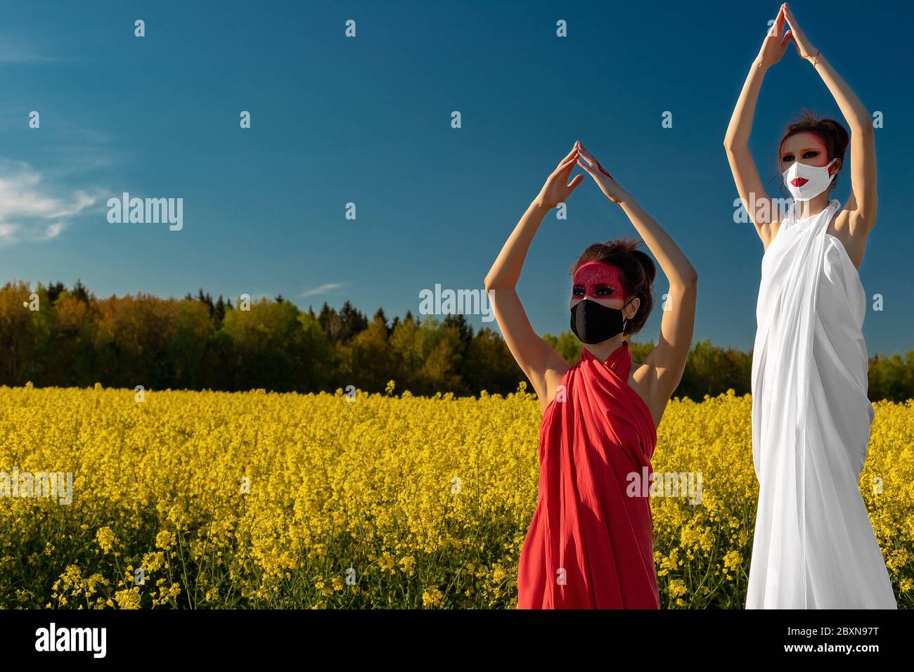 Two beautiful young brunette girls with creative bright makeup in tunics on a background of a field of yellow flowers and blue sky. One girl in a mask Stock Photo