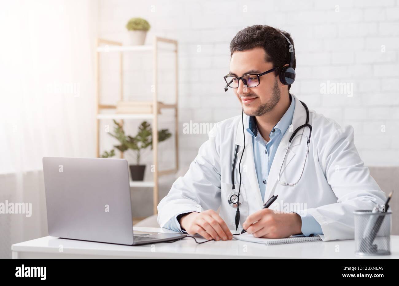 Online lessons and conferences. Doctor makes notes sitting at table with laptop Stock Photo
