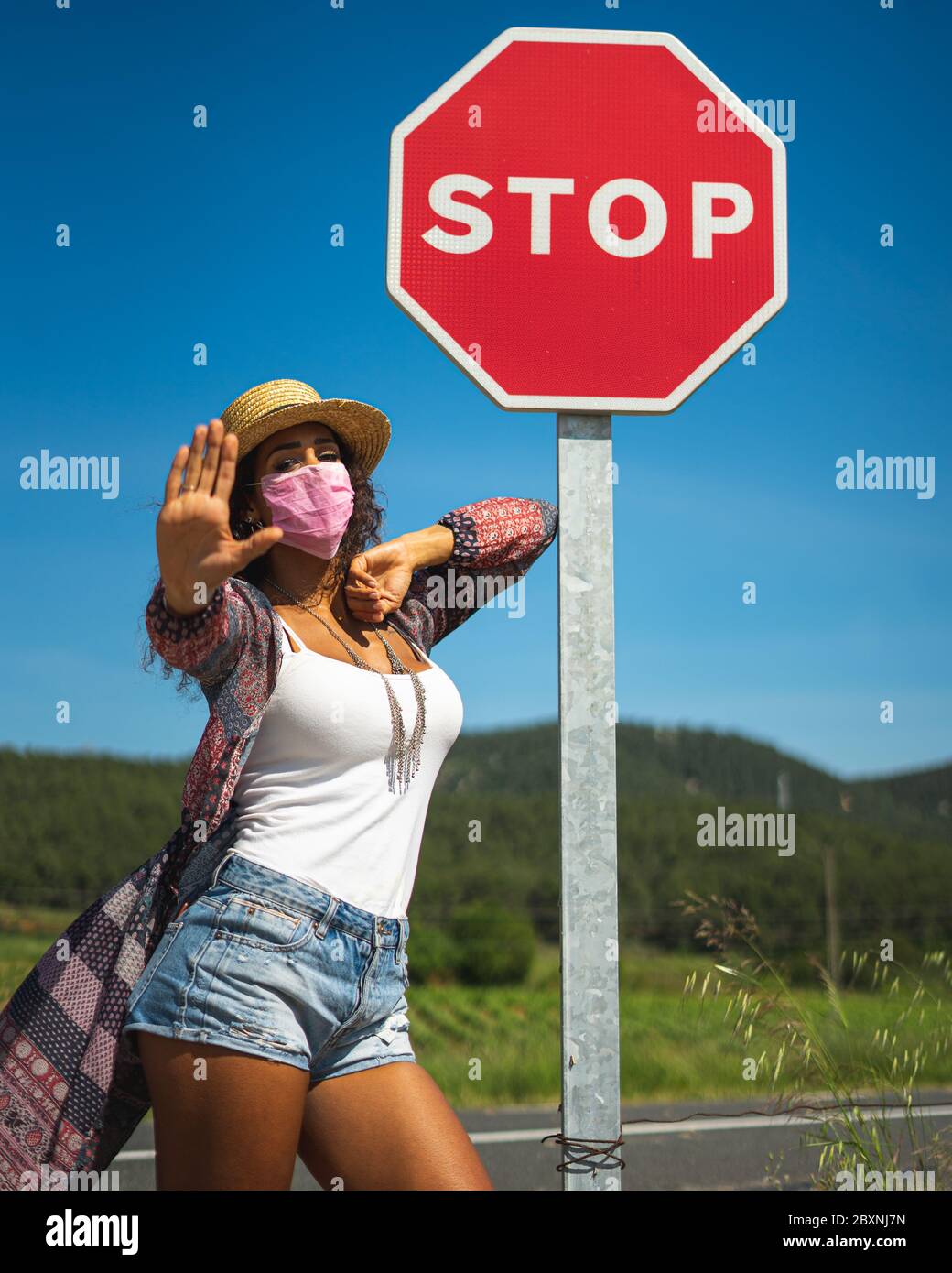 Portrait of a pretty young Brazilian woman looking at the camera with a mask protection for coronavirus infection and a stop sign Stock Photo