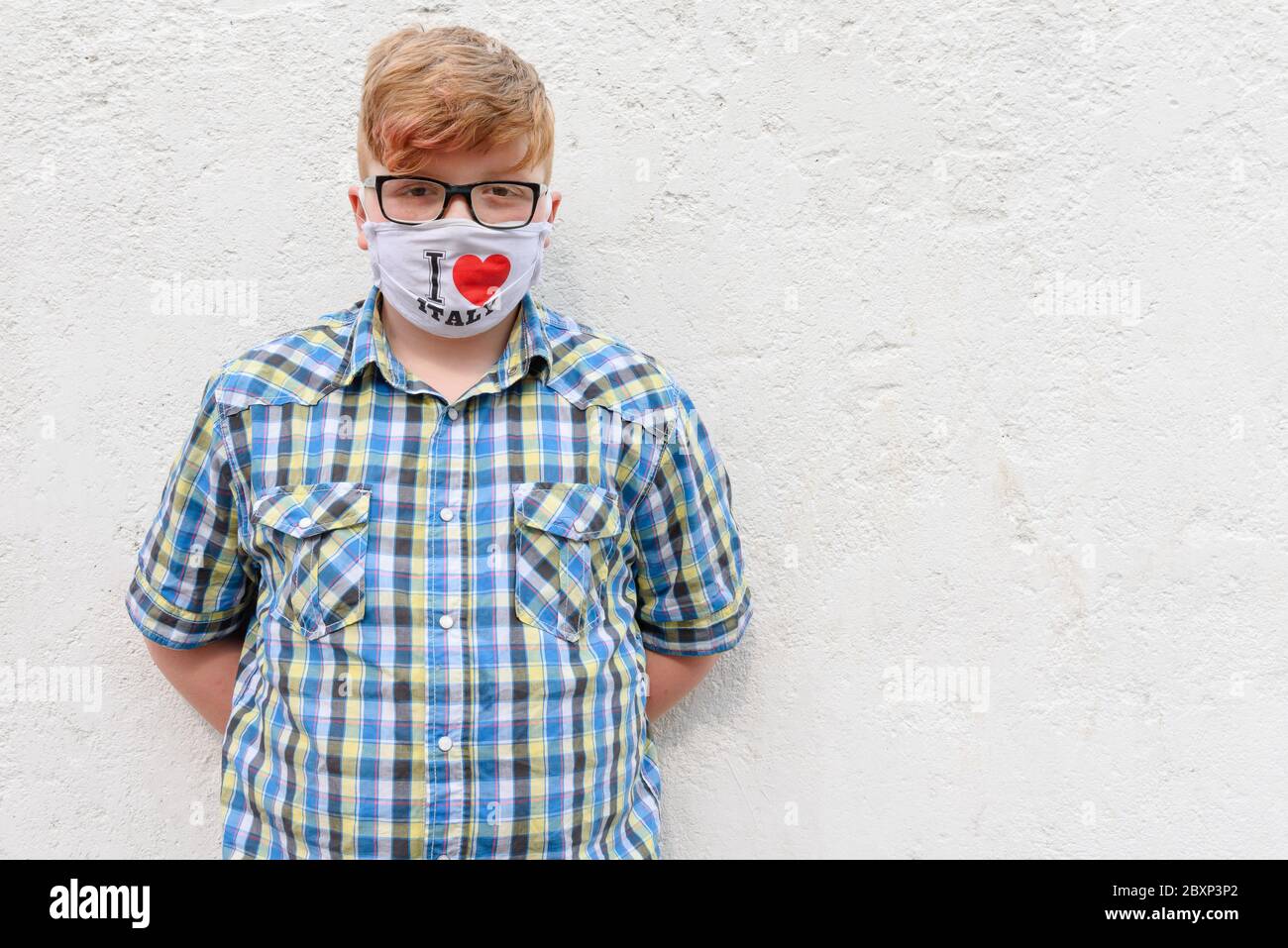 Red-haired boy with glasses and surgical mask with the inscription I LOVE ITALY. A boy in a plaid shirt stands on a wall background. Stock Photo