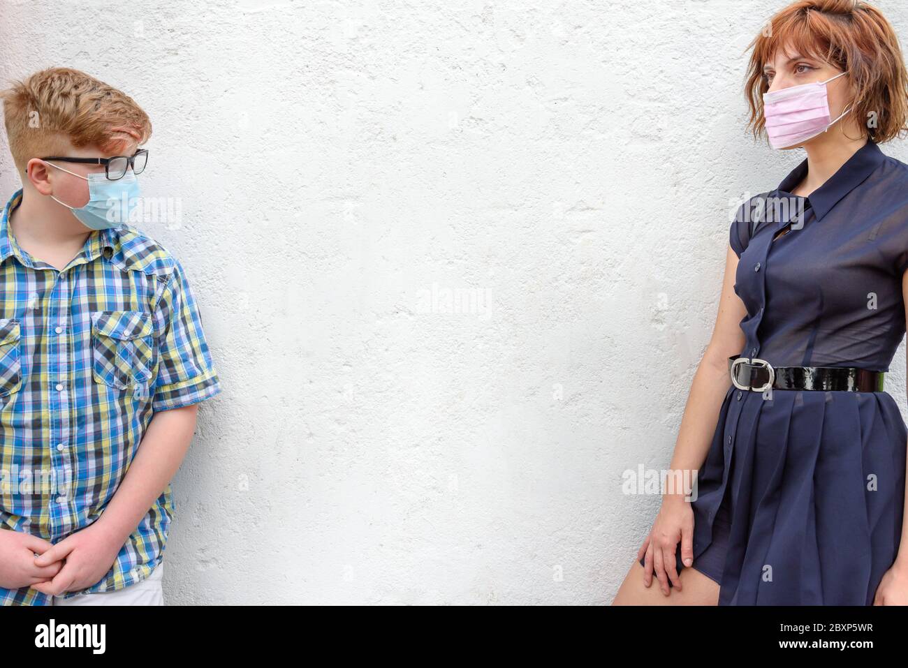 Young red-haired woman and boy wearing a surgical mask stand against a wall background. Sister with trendy glasses and little brother with glasses wea Stock Photo