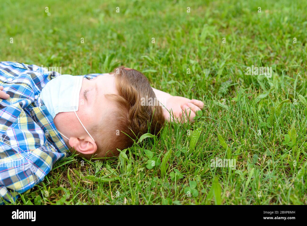 Portrait of a young boy with surgical mask lying on the grass in the park. Close-up of a little boy in a plaid shirt relaxing on the grass. Stock Photo