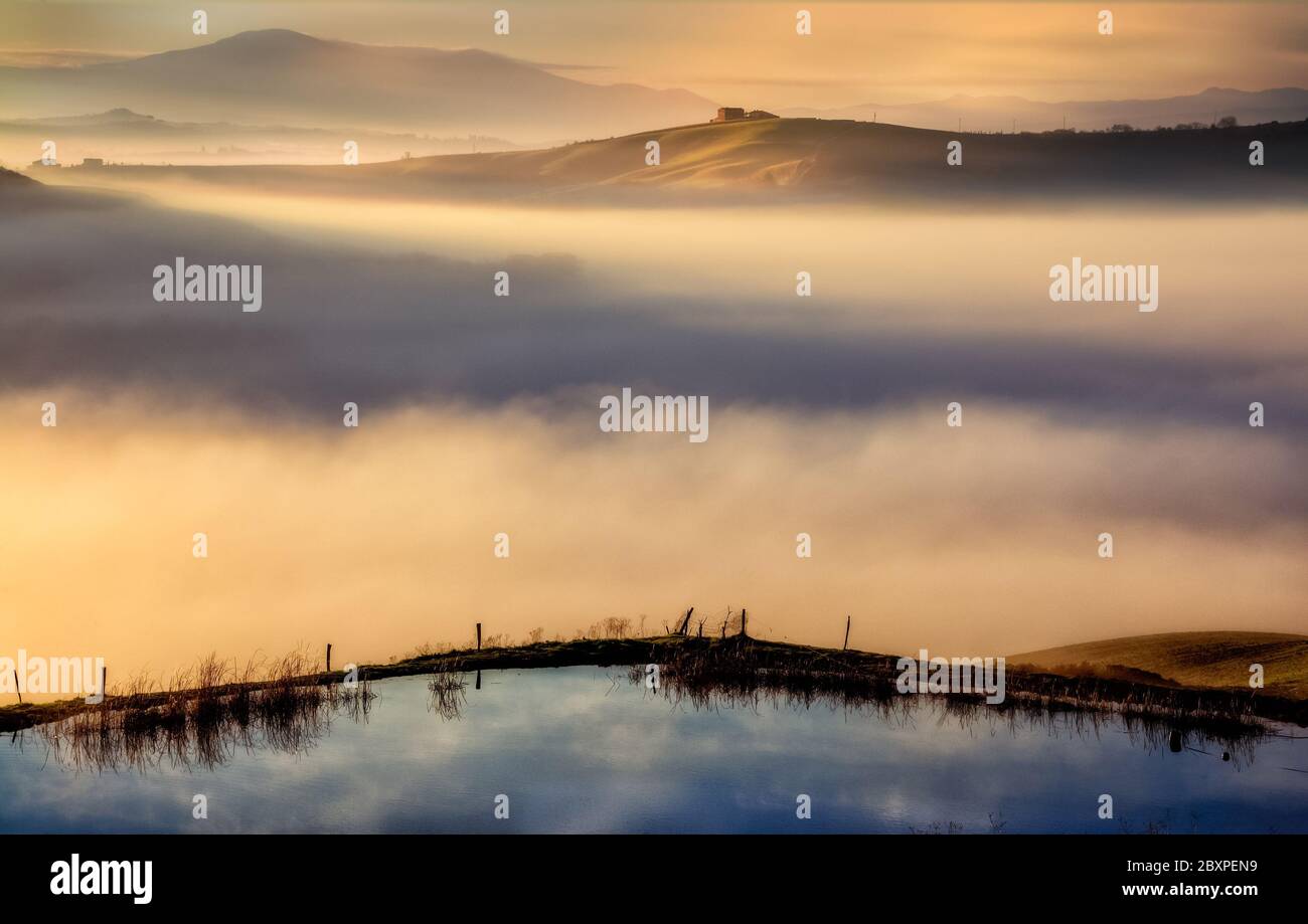 a misty sunrise in the crete senesi area with a little lake in the foreground and a lone farmahouse in the distance Stock Photo
