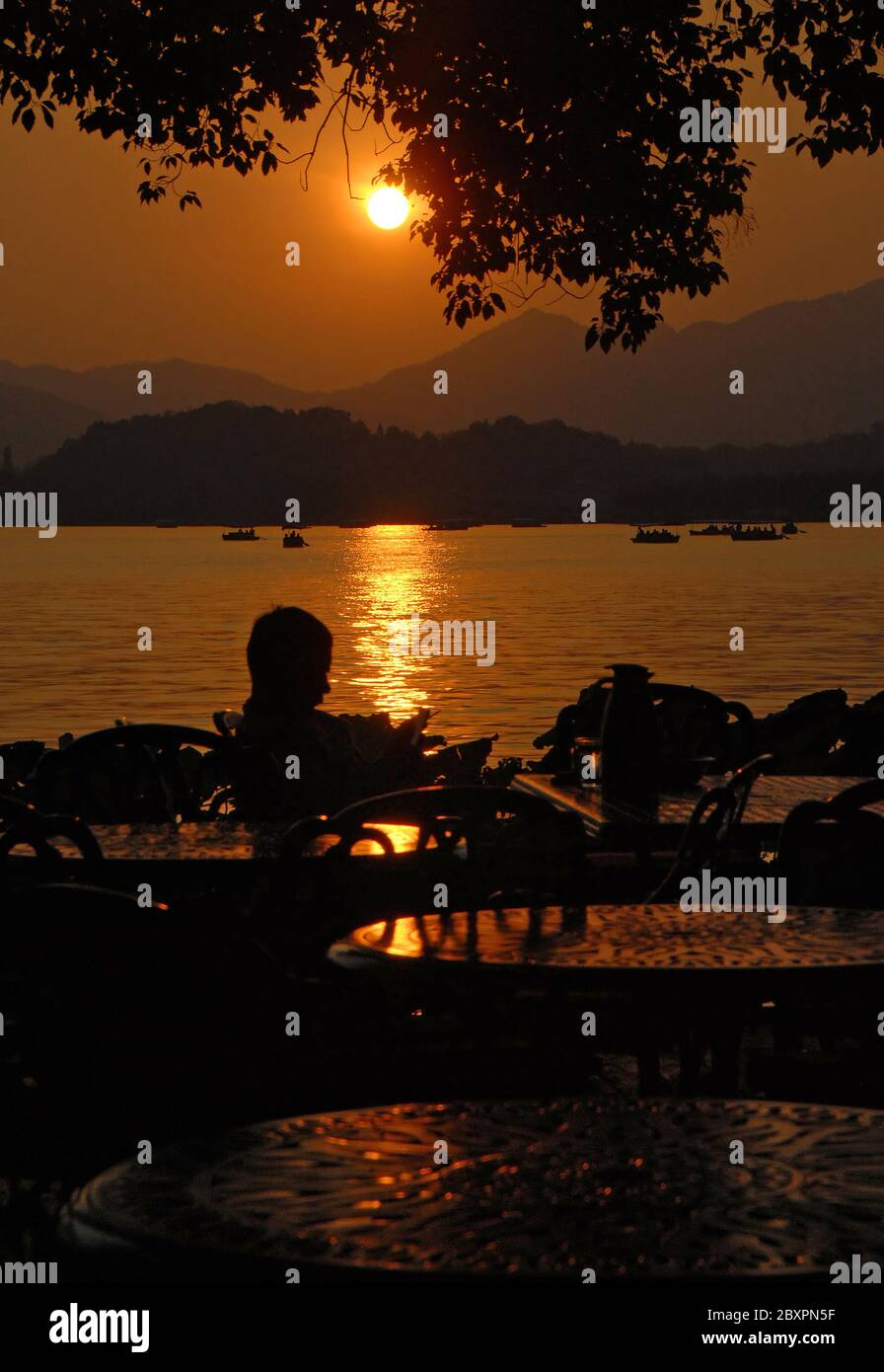 West Lake (Xi Hu) sunset in Hangzhou, Zhejiang Province, China. Sunset view of West Lake, Hangzhou with silhouette of a person sitting in a cafe. Stock Photo